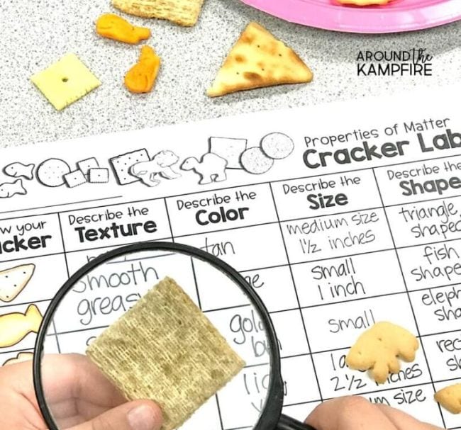 Child's hand holding magnifying glass over a cracker, with worksheet labeled Cracker Lab in the background