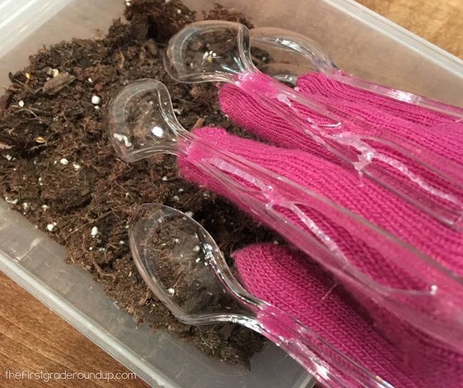 Student's hand wearing pink glove with plastic spoons attached to fingers, digging in dirt