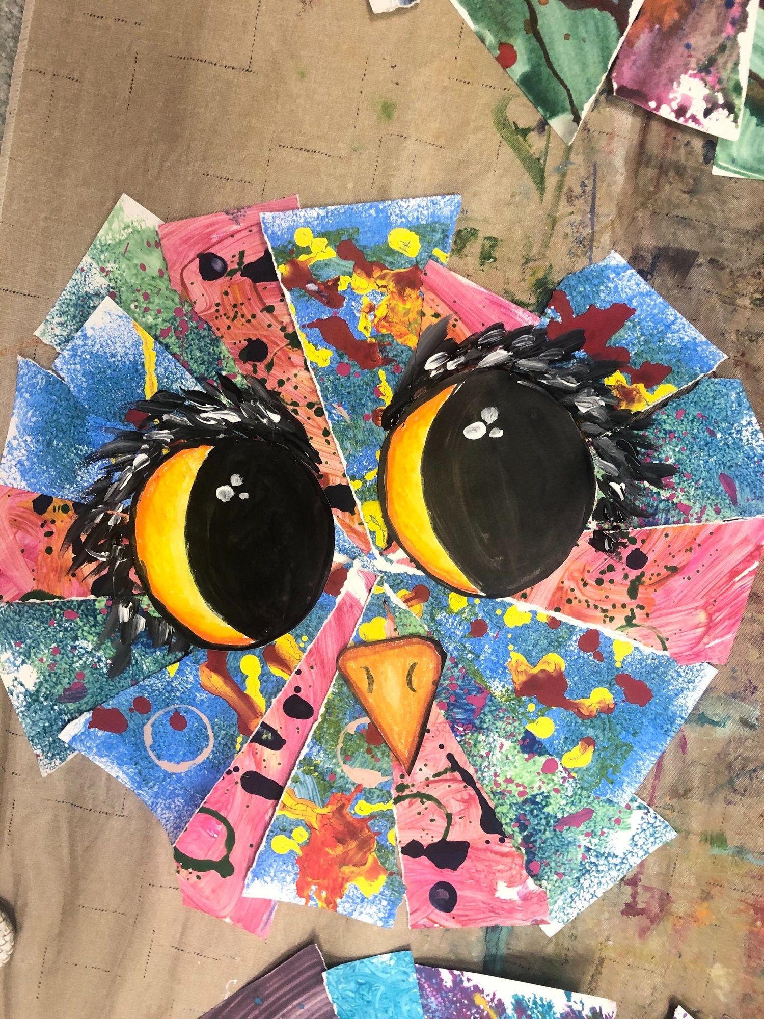 An owl is created from triangluar shaped papers that have paint splattered on them. Two large owl eyes are drawn on and a beak.