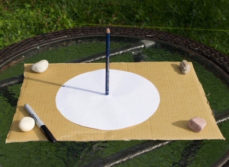 A large piece of cardboard has a white circle in the center with a pencil standing upright in the middle of the circle. Rocks are on all four corners holding it down. 