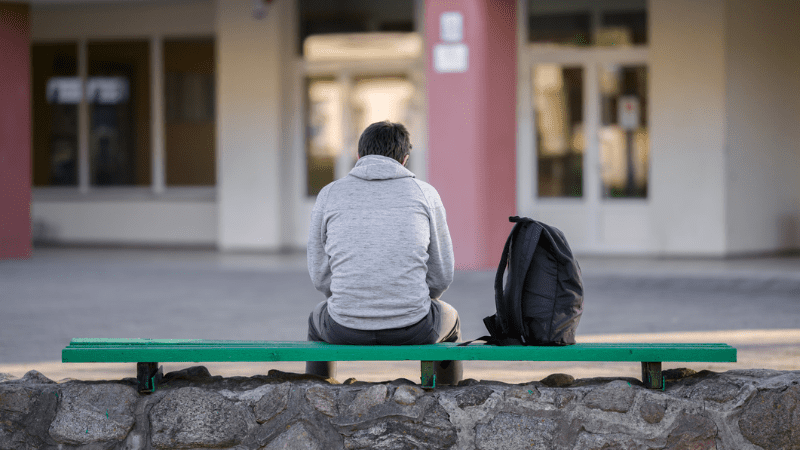 Back of a student alone on a bench with a backpack