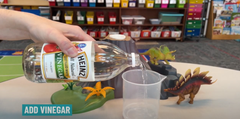 A hand is shown pouring vinegar into a plastic measuring cup in this step of a baking soda volcano.